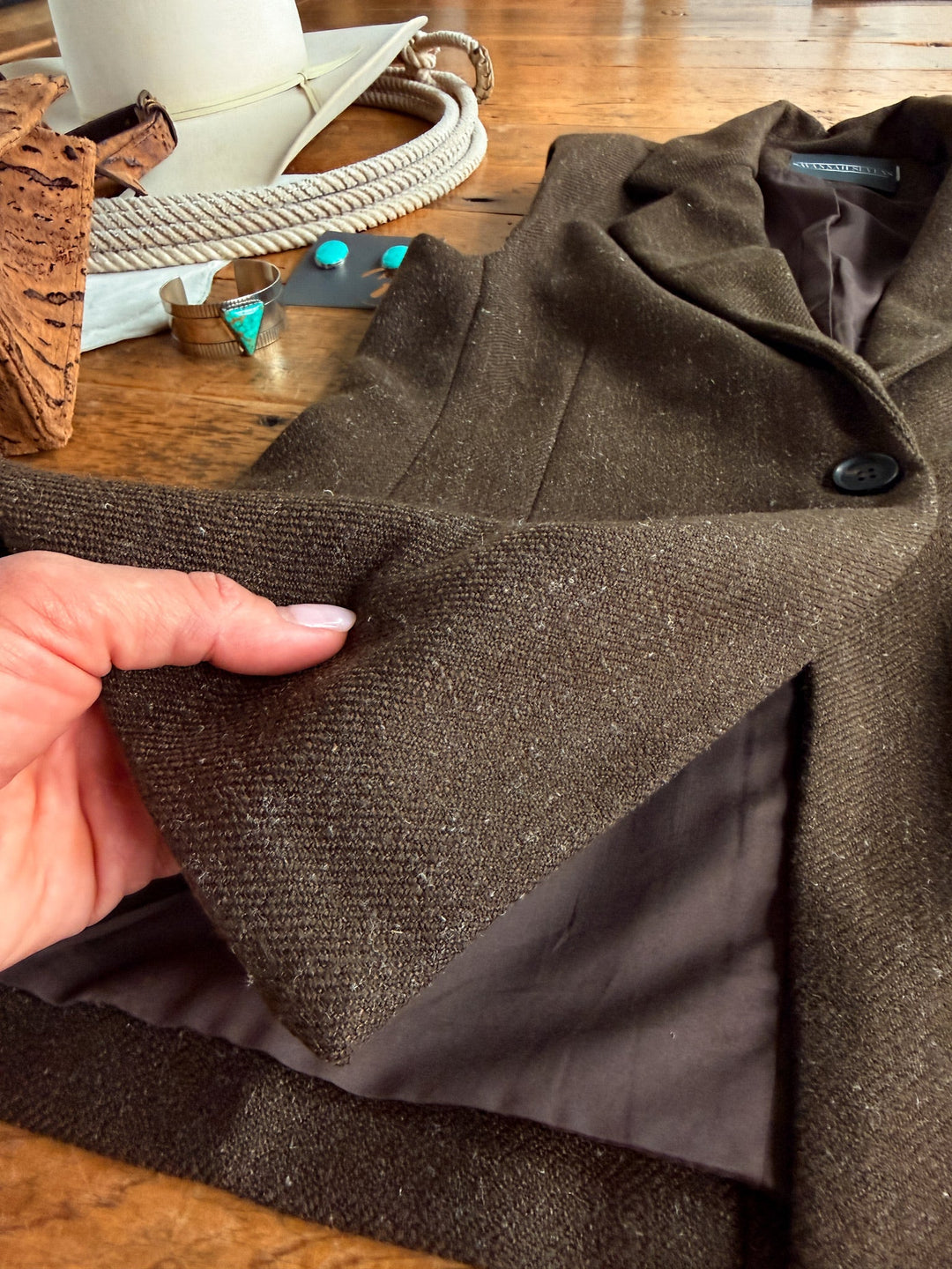 A hand lifts the hem of a brown textured blazer, revealing its dark inner lining. The blazer rests on a wooden table alongside jewelry, a hat, and what appears to be part of the Armona Chocolate Twill Vest Set by Savannah Sevens western life{&}style in the background.