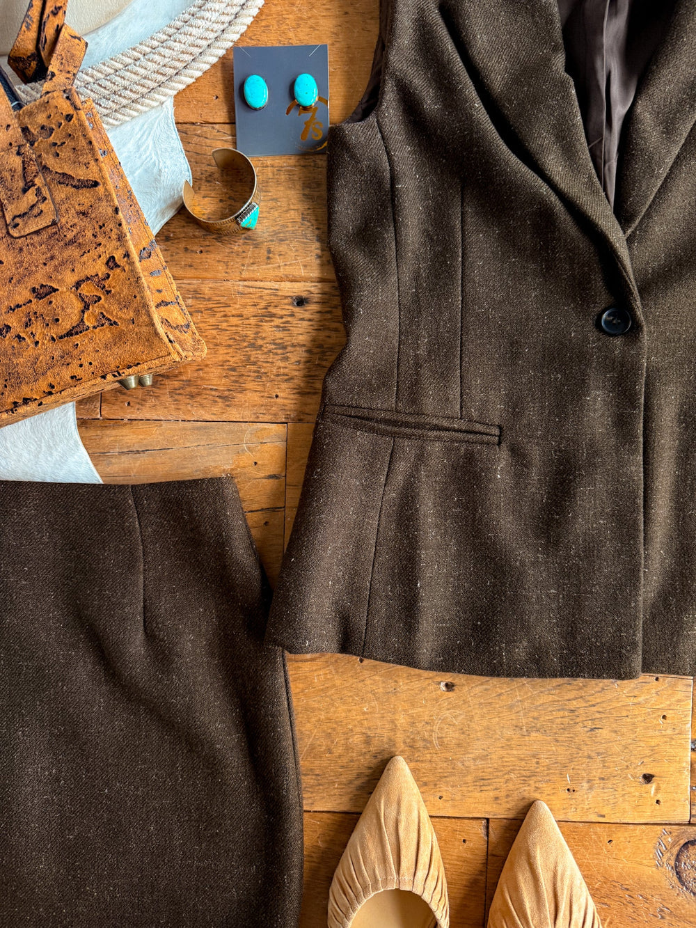 The Armona Chocolate Twill Vest Set by Savannah Sevens western life{&}style, featuring a high-waisted skirt and coordinating single-button vest, is styled with tan pointed-toe flats, a cork handbag, turquoise jewelry, and a rope accessory—all artfully arranged on a wooden surface.