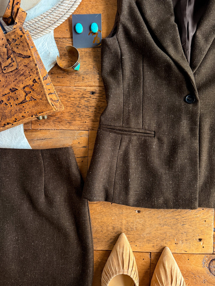 The Armona Chocolate Twill Vest Set by Savannah Sevens western life{&}style, featuring a high-waisted skirt and coordinating single-button vest, is styled with tan pointed-toe flats, a cork handbag, turquoise jewelry, and a rope accessory—all artfully arranged on a wooden surface.