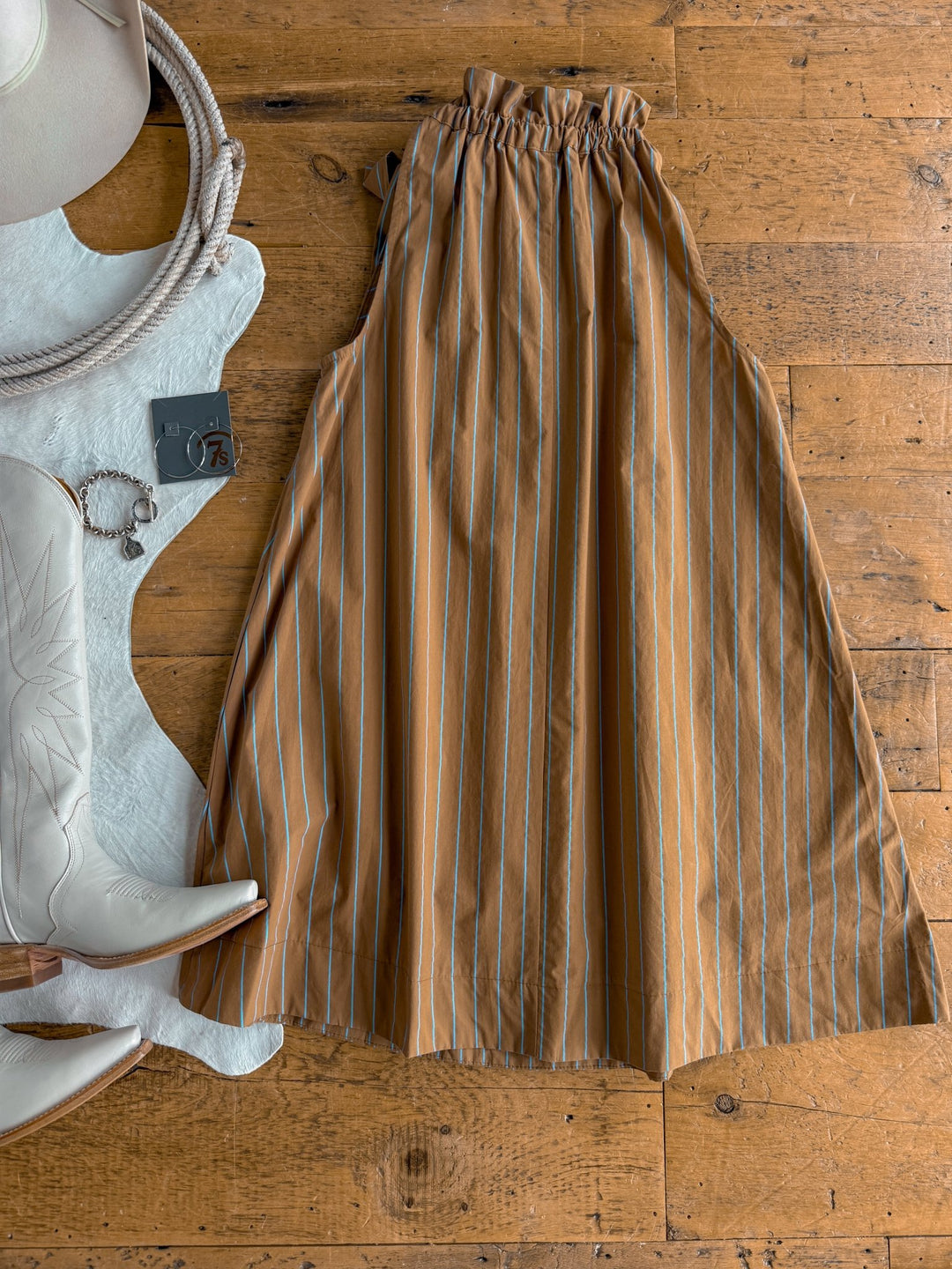The Ashmore Brown Striped Halter Neck Dress by Savannah Sevens western life{&}style is laid flat on a wooden floor next to white cowboy boots, a white hat, a coiled rope, a cowhide rug, and a silver watch with a charm.