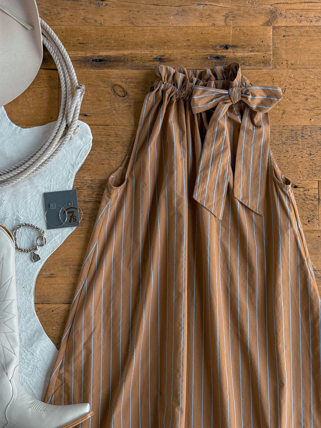 The Ashmore Brown Striped Halter Neck Dress by Savannah Sevens western life{&}style is displayed on a wooden floor, styled with a white hat, a camera, jewelry, and matching white cowboy boots.