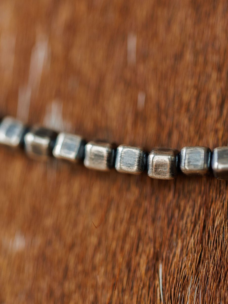 A close-up of the Atoka Necklace by Savannah Sevens western life{&}style, featuring sterling silver square beads, resting on a brown textured surface with the chain in focus and a softly blurred background.