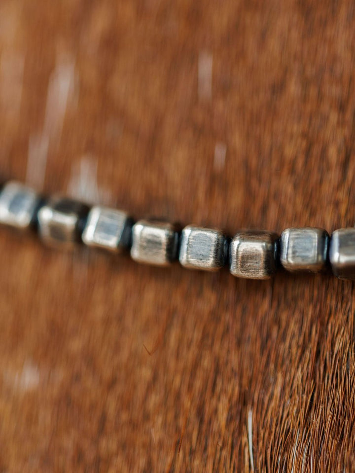 A close-up of the Atoka Necklace by Savannah Sevens western life{&}style, featuring sterling silver square beads, resting on a brown textured surface with the chain in focus and a softly blurred background.