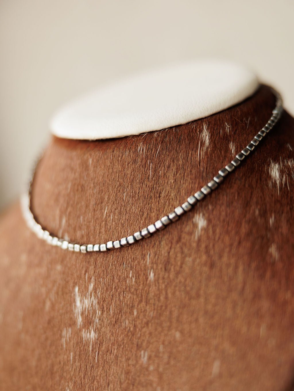 The Atoka Necklace by Savannah Sevens western life{&}style, a sterling silver box chain, is displayed on a brown cowhide-textured jewelry bust with white patches and a white top.