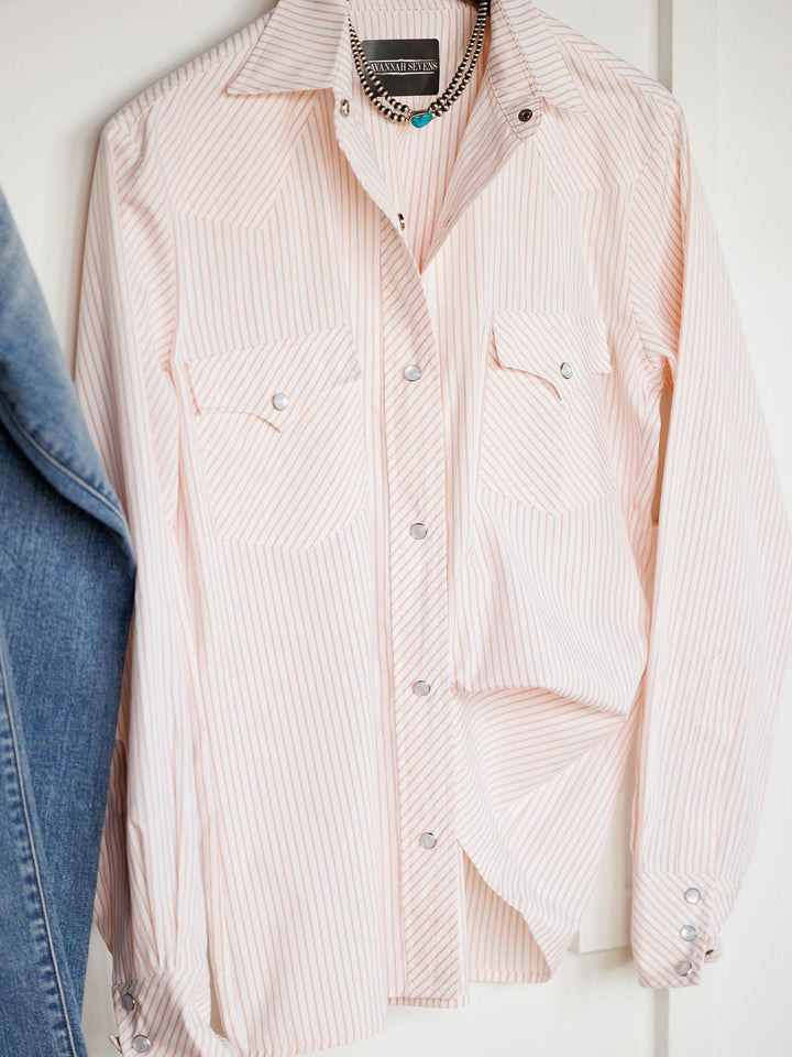 The Savannah Sevens western life{&}style Aullville Striped Pearl Snap Top, featuring light pink and white stripes, snap buttons, and two front pockets, hangs on a hanger beside blue jeans with a silver chain necklace draped over its collar.