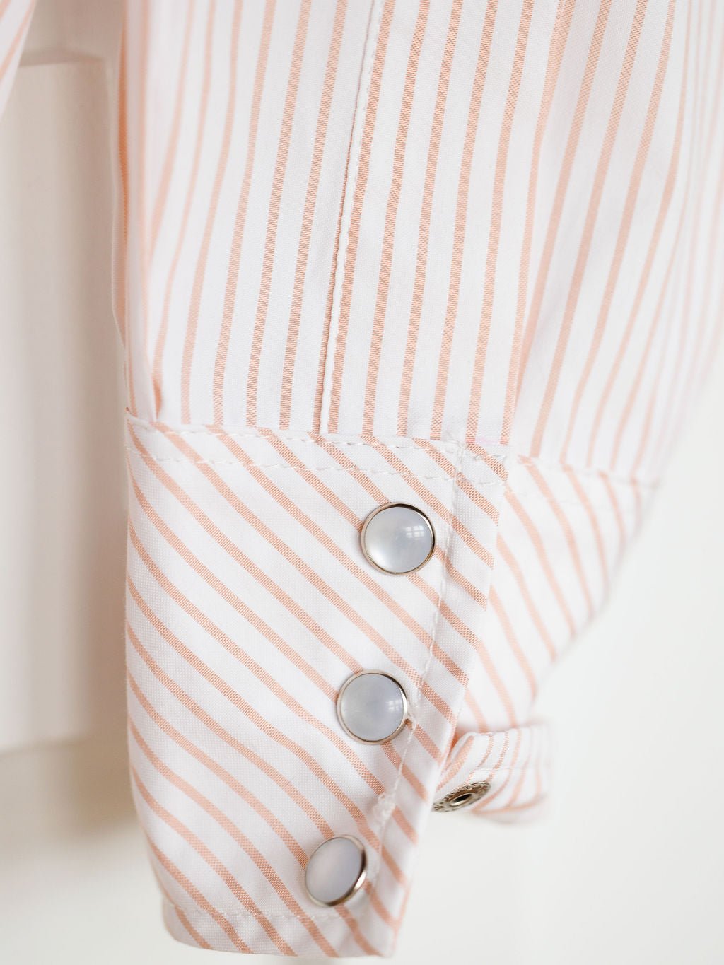 Close-up of the Aullville Striped Pearl Snap Top by Savannah Sevens western life{&}style, featuring a snap button cuff with light pink and white vertical stripes, three shiny pearl snaps, and a diagonally patterned cuff for a unique retro western look.