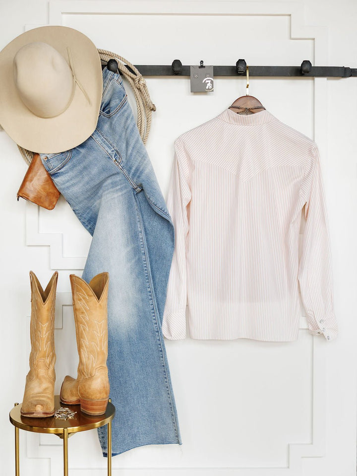 A beige cowboy hat, blue jeans, a Savannah Sevens western life{&}style Aullville Striped Pearl Snap Top, tan cowboy boots, a brown belt, and a rope are arranged neatly against a white wall with decorative molding.
