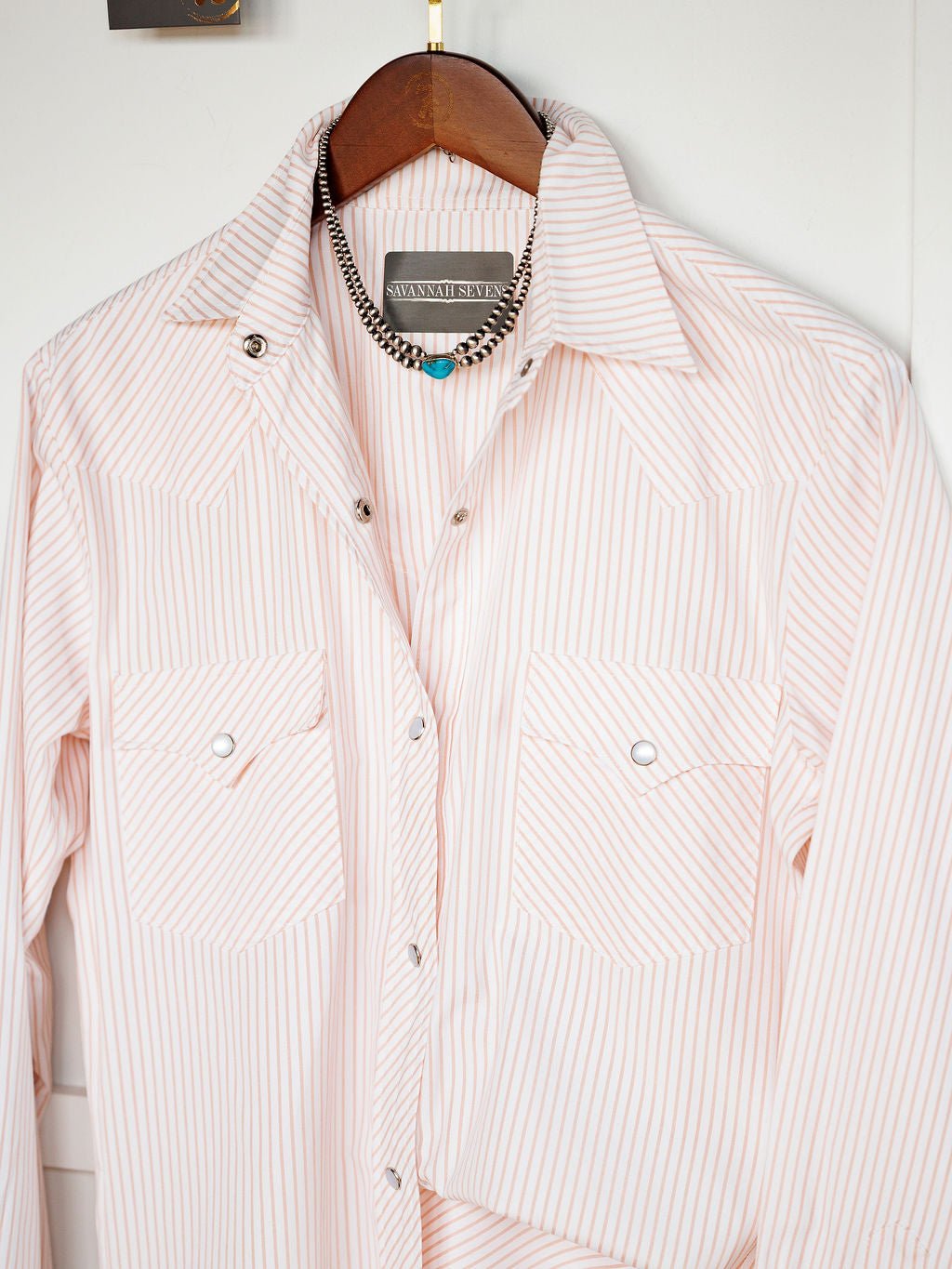 The Aullville Striped Pearl Snap Top by Savannah Sevens western life{&}style, in light pink and white stripes, hangs on a wooden hanger with a silver necklace featuring turquoise and black beads draped above it.