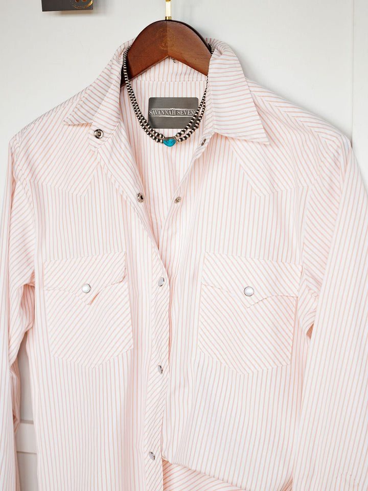 The Aullville Striped Pearl Snap Top by Savannah Sevens western life{&}style, in light pink and white stripes, hangs on a wooden hanger with a silver necklace featuring turquoise and black beads draped above it.