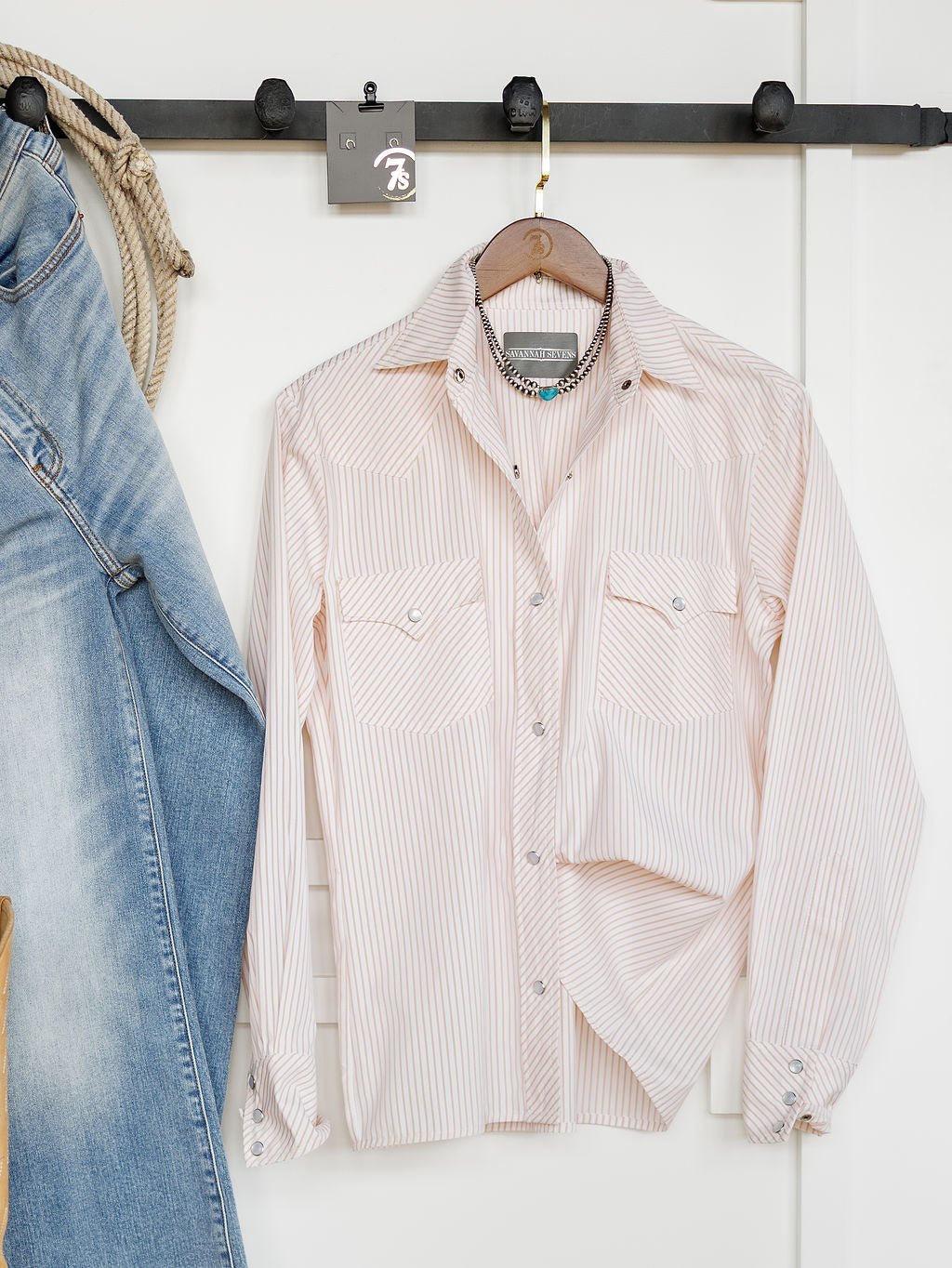 The Savannah Sevens western life{&}style Aullville Striped Pearl Snap Top in light pink and white stripes hangs on a wooden hanger from a black wall-mounted rack, beside blue jeans and a coiled rope.