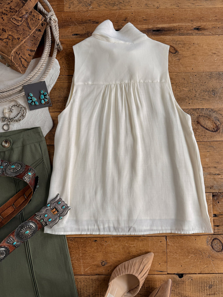 The Aurora Cream Sleeveless Top by Savannah Sevens western life{&}style is laid flat on a wooden surface, surrounded by a green skirt, turquoise jewelry, a brown belt, tan heels, cork pads, and other accessories. This blouse features an adjustable tie neckline for added versatility.