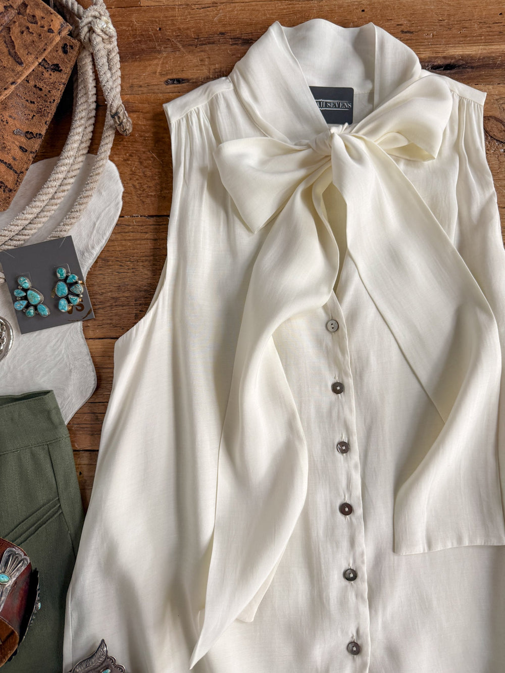 The Aurora Cream Sleeveless Top by Savannah Sevens western life{&}style is a white, sleeveless button-up featuring a large adjustable tie neckline bow, button-down front, and gathered shoulders. Shown styled for young contemporary sizing and laid on a wooden surface beside turquoise earrings and green pants.