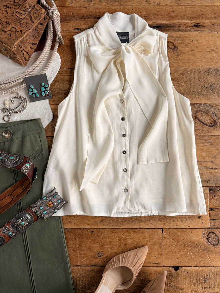 Wearing the Aurora Cream Sleeveless Top by Savannah Sevens western life{&}style, styled with sage green pants, tan heels, a brown belt, turquoise jewelry, and a brown purse—all set against a wooden floor.