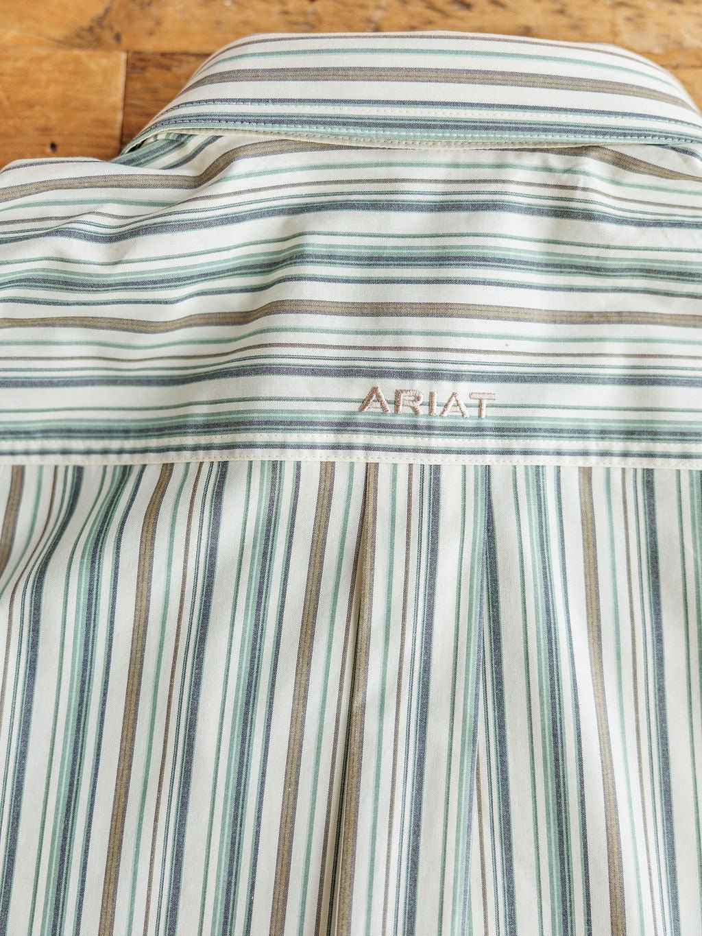 A close-up of the Austwell Ariat Men's Pinstripe Button Up by Savannah Sevens western life{&}style shows green, blue, and beige stripes with Ariat embroidery near the collar, laid on a wooden surface.