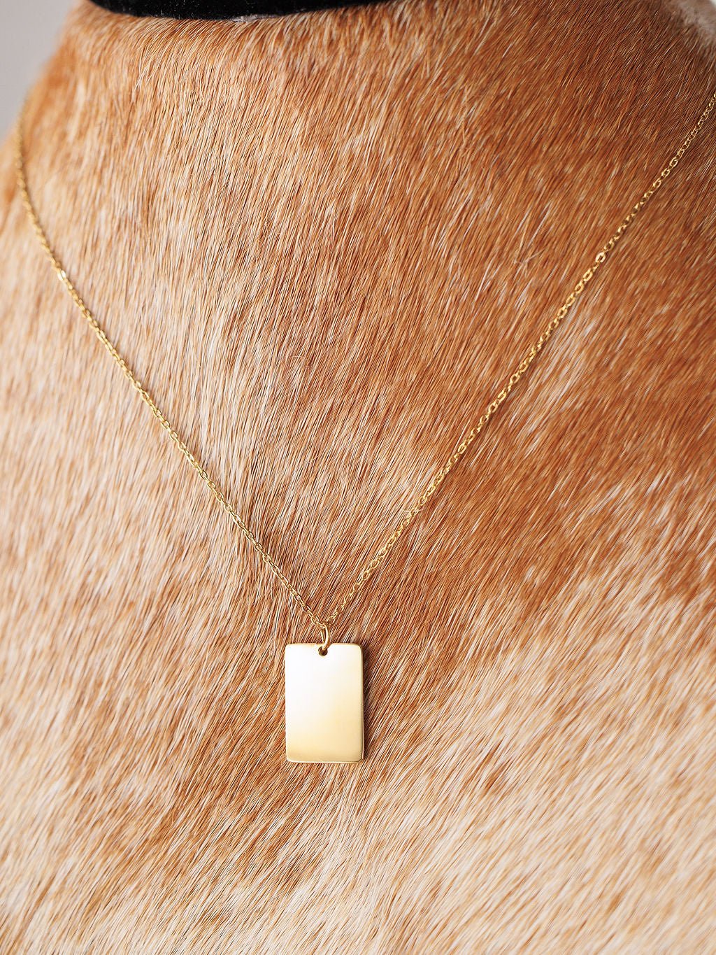 A close-up of the Avera Necklace by Savannah Sevens western life{&}style, an 18K gold coated, tarnish resistant square pendant, worn around the neck of a light brown cowhide with white spots.