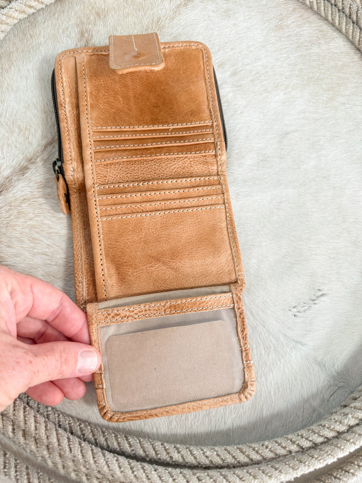 A hand holds open the Avera Oat Leather Snap Wallet by Savannah Sevens western life{&}style, featuring multiple card slots and a transparent ID window. It rests on a light fur surface with a coiled rope nearby, blending stylish design with everyday functionality.
