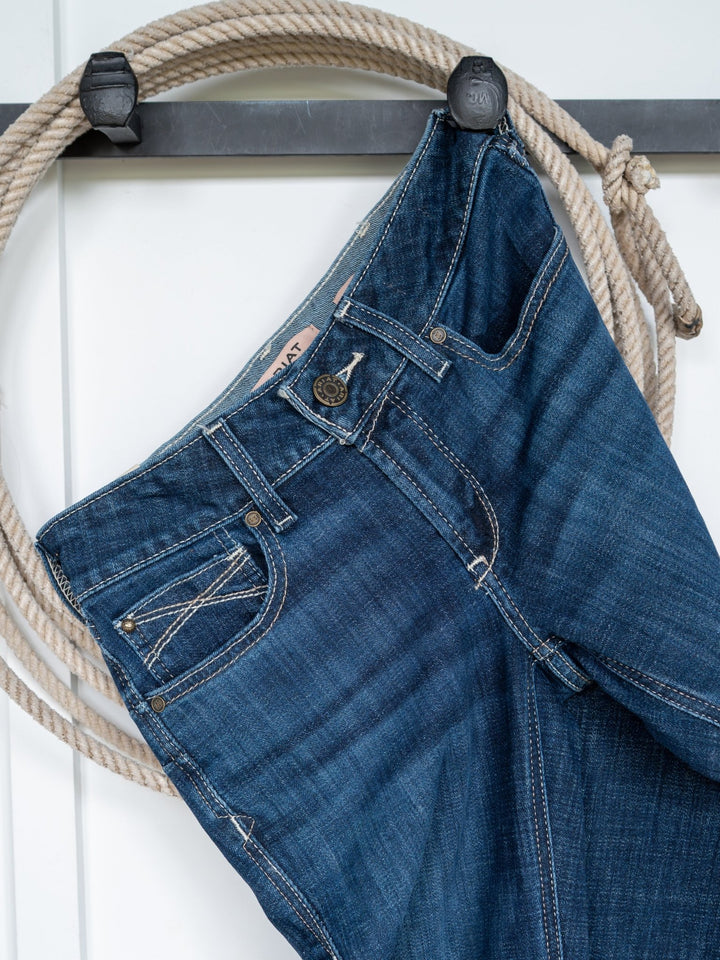 The Avery Perfect Rise Boot Cut Jean by Savannah Sevens western life{&}style in medium dark wash hangs by a belt loop on a black hook, with a coiled beige rope behind it against a white background.
