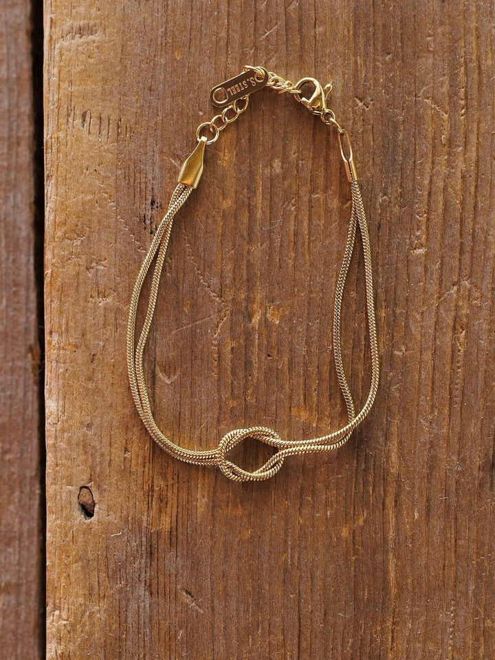 The Aviston Bracelet by Savannah Sevens western life{&}style is a delicate, hypoallergenic gold piece with a small knot detail, thin shiny chain, lobster clasp, and a small charm near the clasp, displayed on rustic wood.