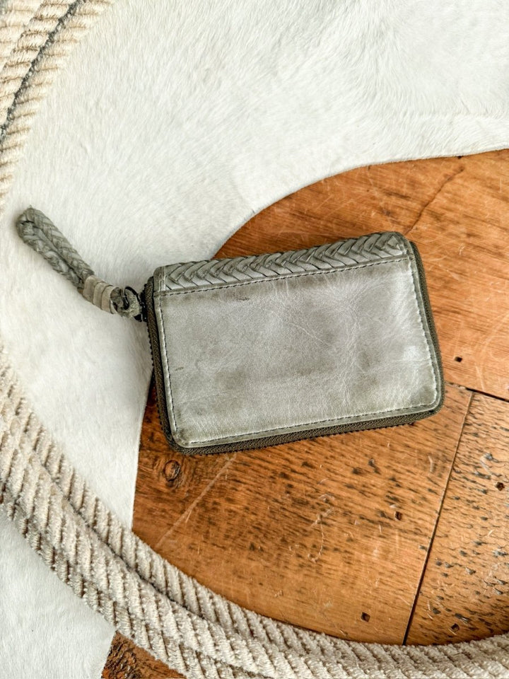The Avoca Wallet by Savannah Sevens western life{&}style is a small, light gray handcrafted wallet with braided details, displayed on a wooden surface near a coiled rope and a white textured background.