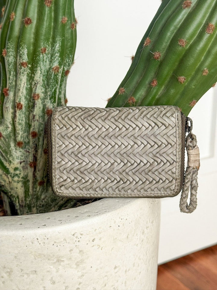 The Avoca Wallet by Savannah Sevens western life{&}style, a small light gray handcrafted piece with a braided zipper pull, is displayed against a large green cactus in a white textured pot.