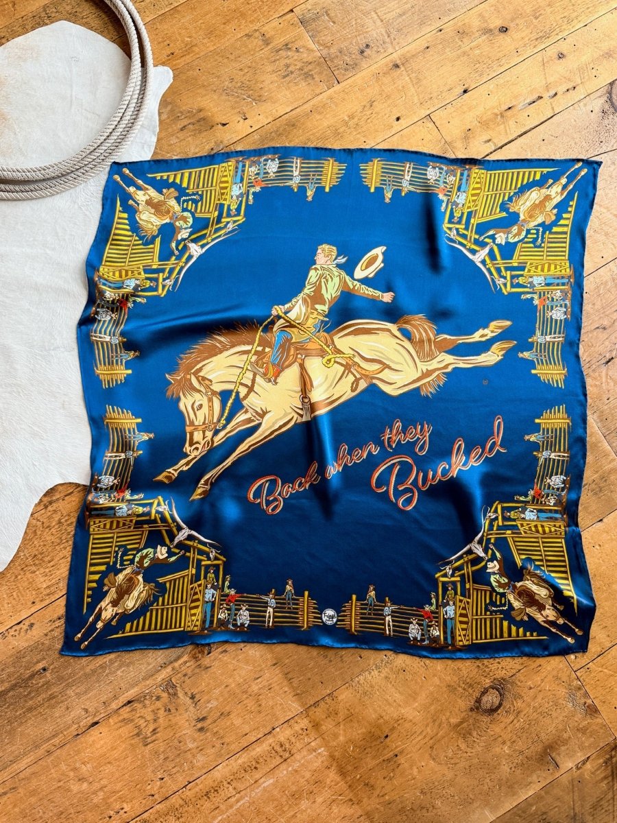 The Back When Wild Rag by Savannah Sevens western life{&}style is a blue silk scarf with cowboy graphics, “Back when they Bucked” in script, and a gold and brown rodeo scene border. Shown displayed on a wooden floor.