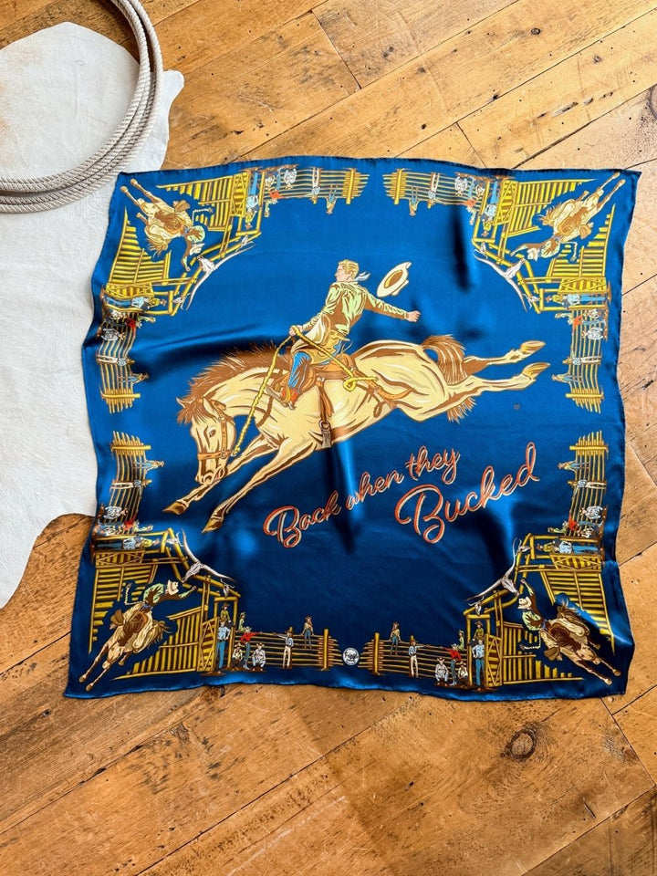 The Back When Wild Rag by Savannah Sevens western life{&}style is a blue silk scarf with cowboy graphics, “Back when they Bucked” in script, and a gold and brown rodeo scene border. Shown displayed on a wooden floor.