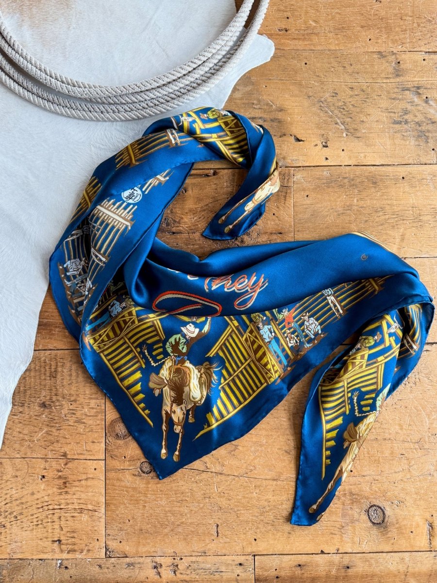 The Back When Wild Rag by Savannah Sevens western life{&}style—a blue silk scarf with yellow and gold cowboy graphics—rests folded on a wooden surface beside a coiled rope and pale fabric.