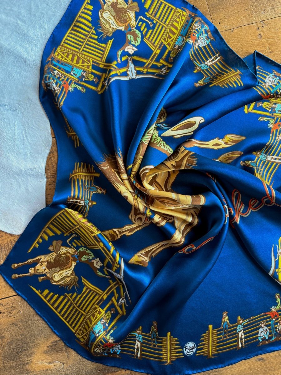 The Back When Wild Rag by Savannah Sevens western life{&}style features intricate cowboy graphics and golden-brown details on silky blue fabric, making its retro rodeo style and vibrant colors stand out on any wooden surface.