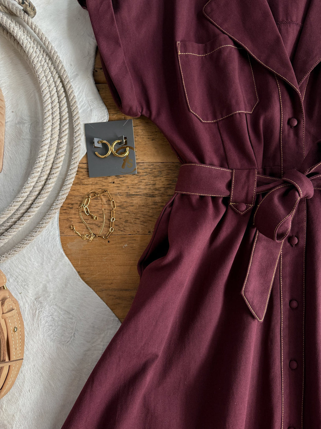The Barada Wine Button Up Dress by Savannah Sevens western life{&}style, featuring short sleeves and tan stitching in a rich burgundy hue, is displayed alongside gold jewelry, tan sandals, and a coiled rope on a wood and white textured surface.
