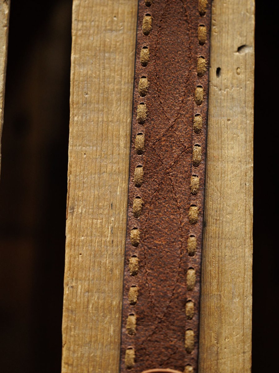 Close-up of the Barlow Belt by Savannah Sevens western life{&}style—a weathered brown leather belt with visible buck stitch detail, shown vertically on a textured, grooved wooden surface.