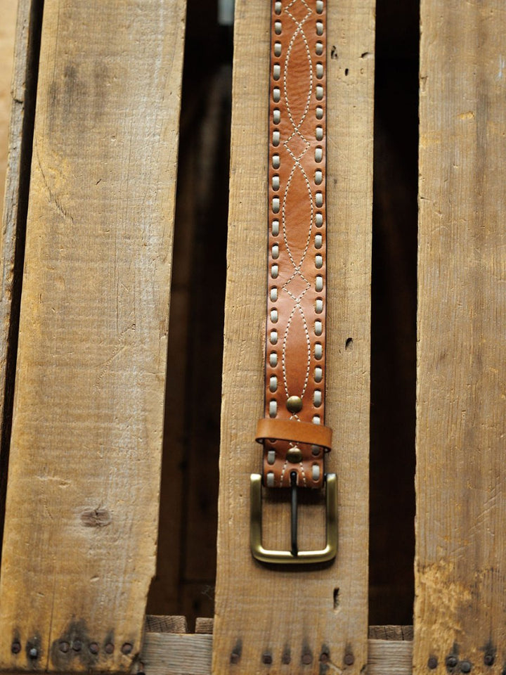 The Barlow Belt by Savannah Sevens western life{&}style, featuring decorative stitching, buck stitch, metal studs, brass buckle, and oval embossed designs, is displayed vertically on a rustic wooden surface.