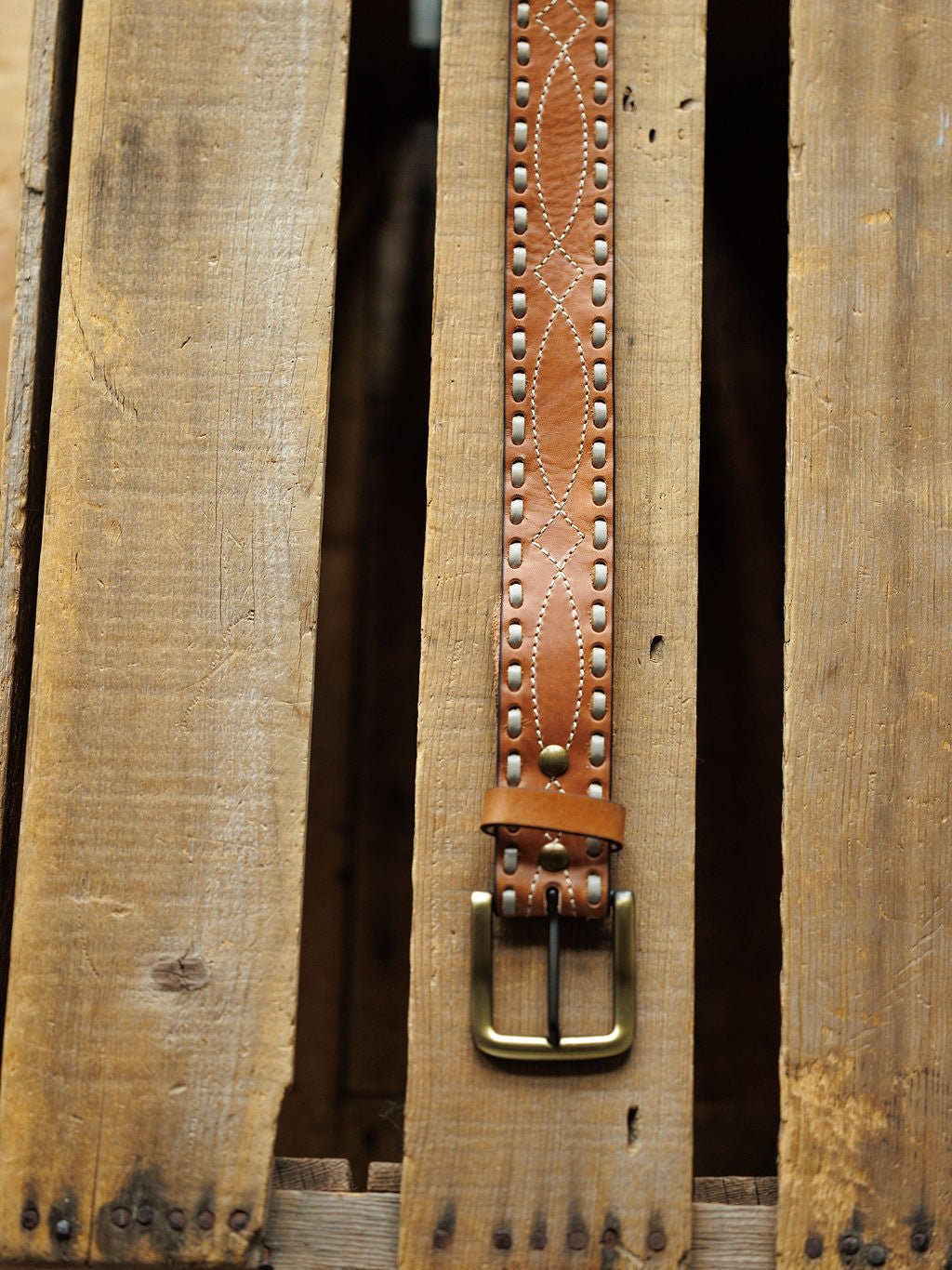 The Barlow Belt by Savannah Sevens western life{&}style, featuring decorative stitching, buck stitch, metal studs, brass buckle, and oval embossed designs, is displayed vertically on a rustic wooden surface.