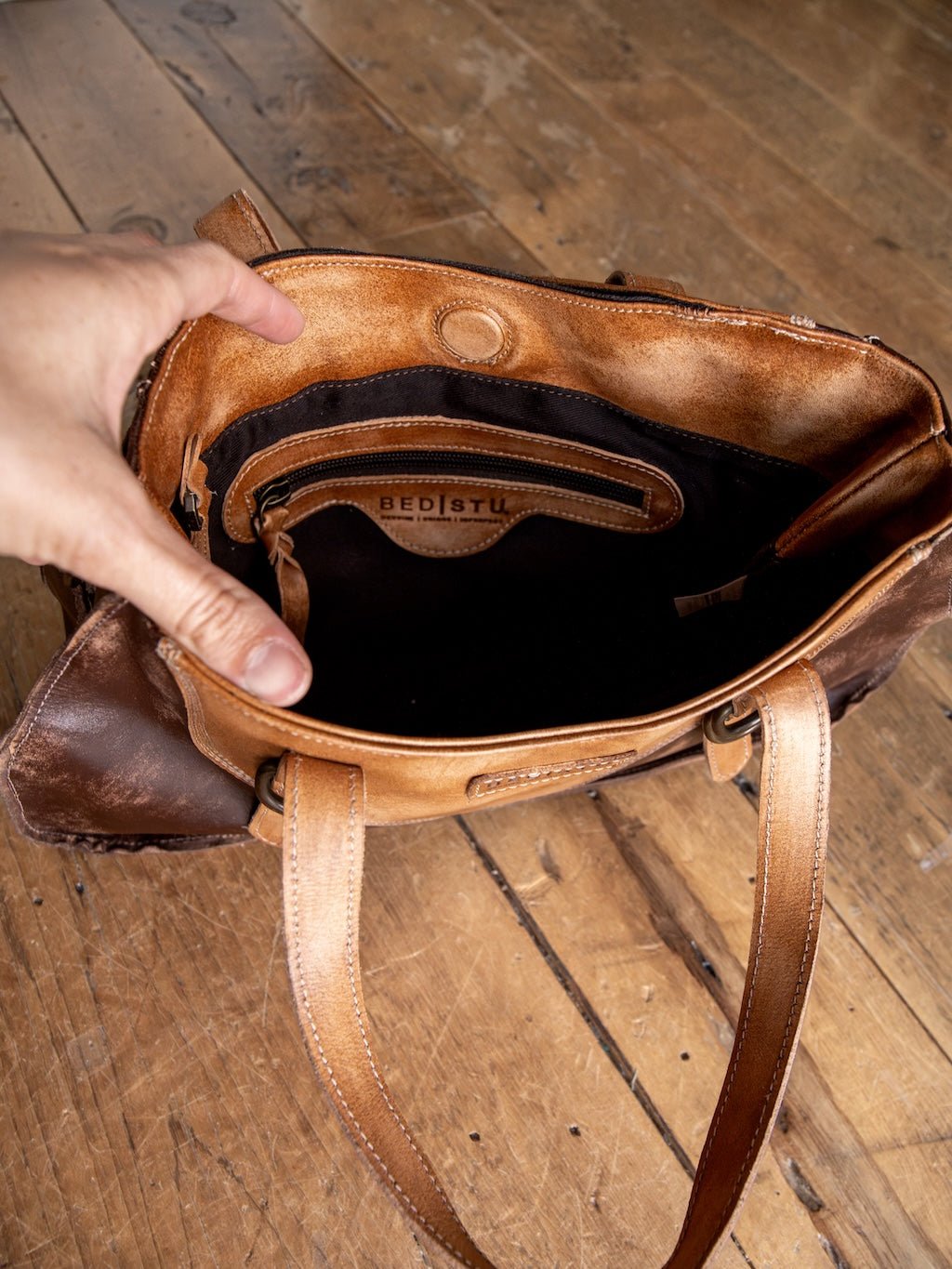 A hand holds open a brown Barstow Leather Handbag by Savannah Sevens western life{&}style, revealing its empty interior and a small inner pocket, placed on a wooden floor.