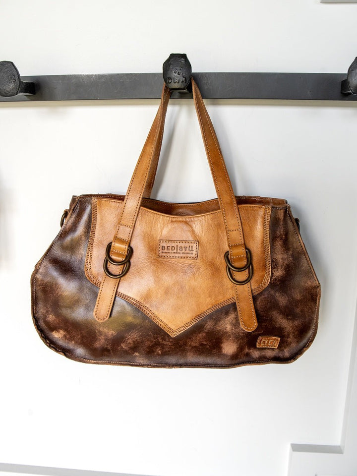 A Barstow Leather Handbag by Savannah Sevens western life{&}style, crafted from two-tone brown distressed leather with double handles, hangs on a black wall hook. The bag features a stitched pattern and metal rings attaching the handles.