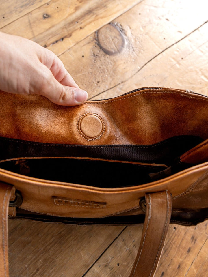 A hand opens the Barstow Leather Handbag by Savannah Sevens western life{&}style, revealing its suede interior and round magnetic snap closure, with the distressed brown leather bag resting on a wooden surface.