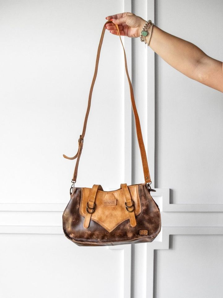 A hand holds a Barstow Leather Handbag by Savannah Sevens western life{&}style, featuring a two-tone brown finish and a long adjustable strap, in front of a white paneled wall.