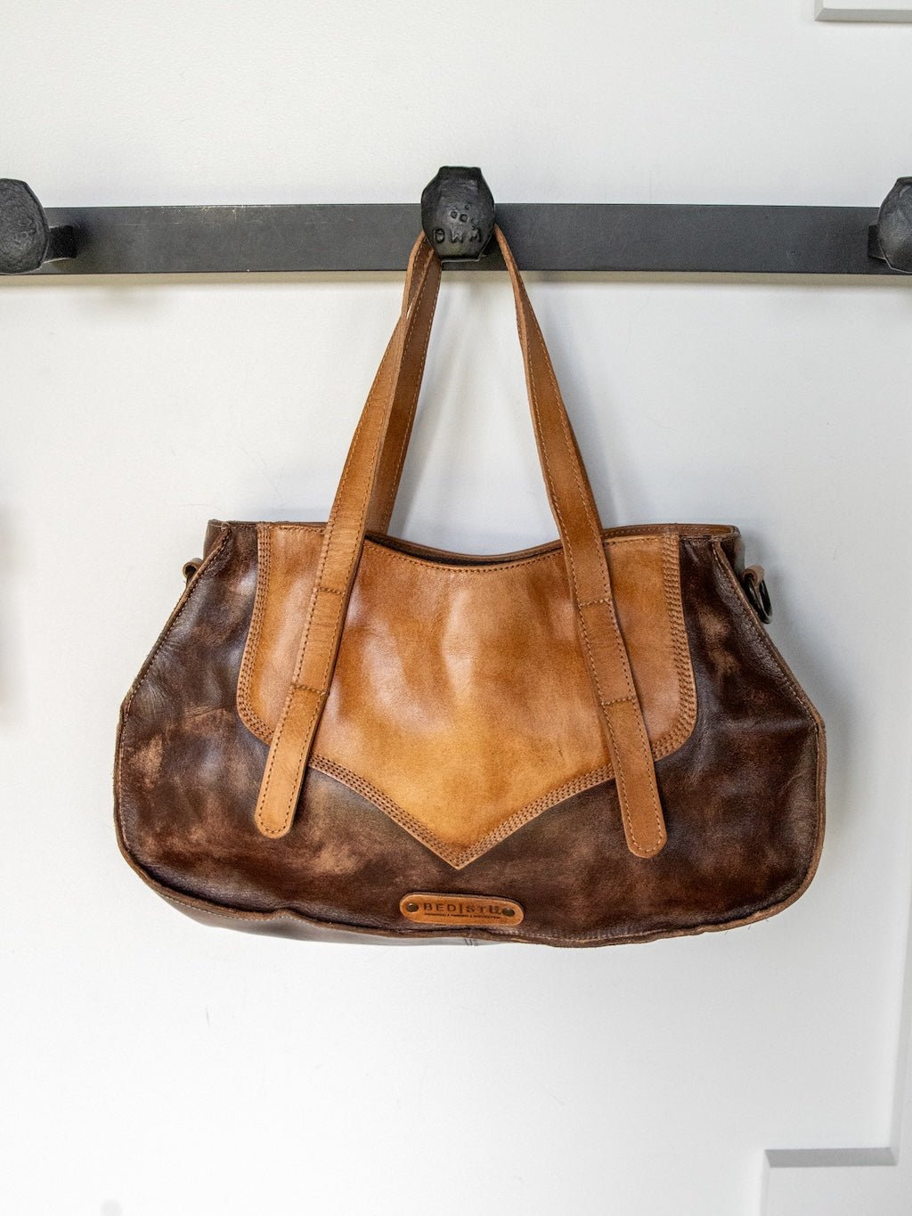 A two-tone brown Barstow Leather Handbag by Savannah Sevens western life{&}style, crafted from distressed leather with dual handles, hangs on a black wall hook against a white background. This western crossbody handbag features detailed stitching and displays a visible brand label at the front bottom center.