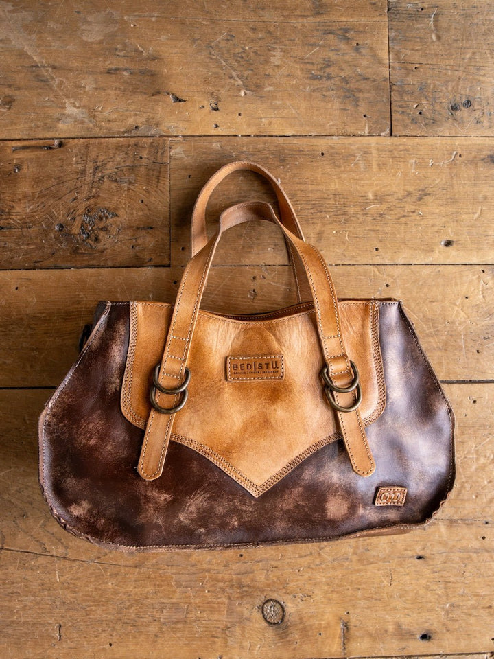 A Barstow Leather Handbag by Savannah Sevens western life{&}style, crafted from two-tone brown distressed leather with dual handles, brass rings, and visible stitching, rests on a rustic wooden floor.