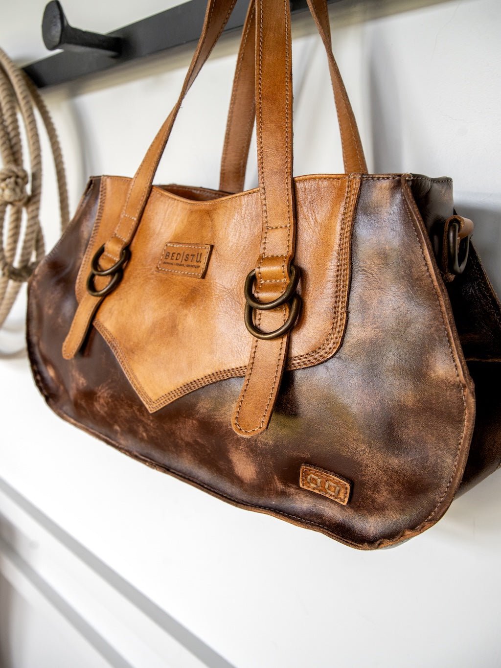 A brown distressed Barstow Leather Handbag by Savannah Sevens western life{&}style, featuring dual handles, metal buckles, and a stitched logo patch, hangs on a wall hook beside a coiled rope—showcasing its signature western leather crossbody style.
