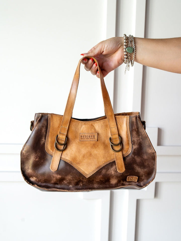 A hand holds the Barstow Leather Handbag by Savannah Sevens western life{&}style, crafted from distressed leather with double handles, metallic rings, and a front label, set against a white background.