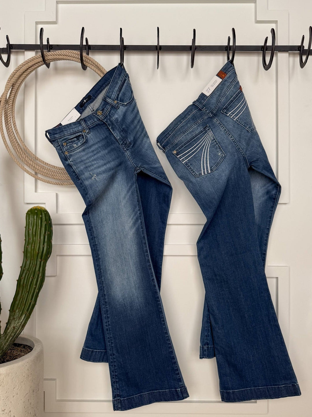 Two pairs of The Beau 7 for all Mankind Dojo Trouser jeans by Savannah Sevens western life{&}style hang side by side on black hooks against a white paneled wall, with a round rope decor and tall cactus in a white pot on the left.