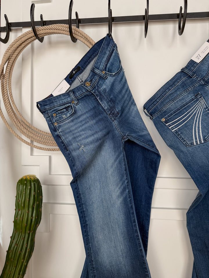 Two pairs of The Beau 7 for all Mankind Dojo Trouser jeans from Savannah Sevens western life{&}style, in medium dark wash with a relaxed fit, hang on black hooks against a white wall, accented by a coiled rope and green cactus.
