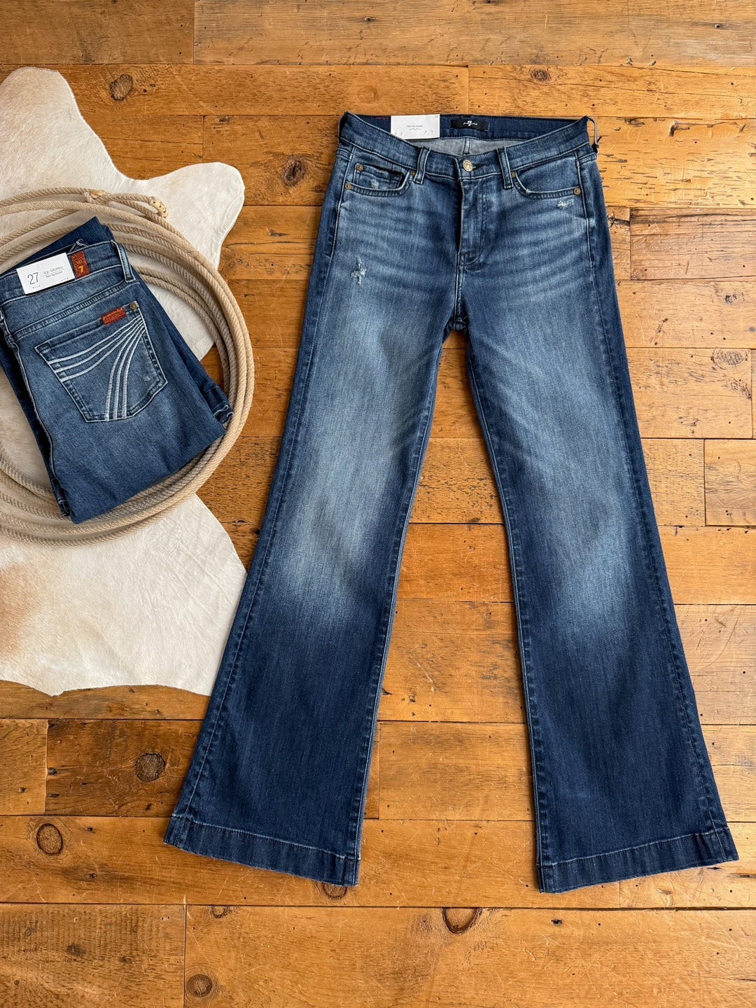 A pair of flared blue jeans in medium dark wash from Savannah Sevens western life{&}style is laid flat on a wooden floor, with two folded pairs nearby, including The Beau 7 for all Mankind Dojo Trouser, by a white rug and coiled rope.