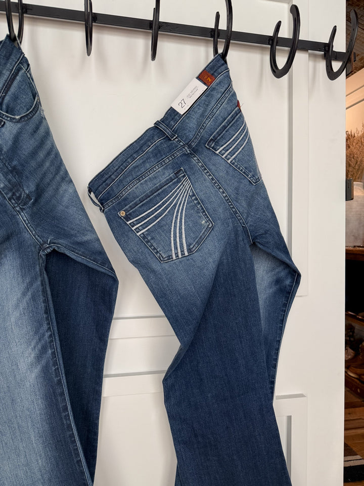 The Beau 7 for all Mankind Dojo Trouser jeans by Savannah Sevens western life{&}style, in a medium dark wash with a relaxed fit, hang by the belt loop on a black hook rack attached to a white door; another pair is partially visible on the left.