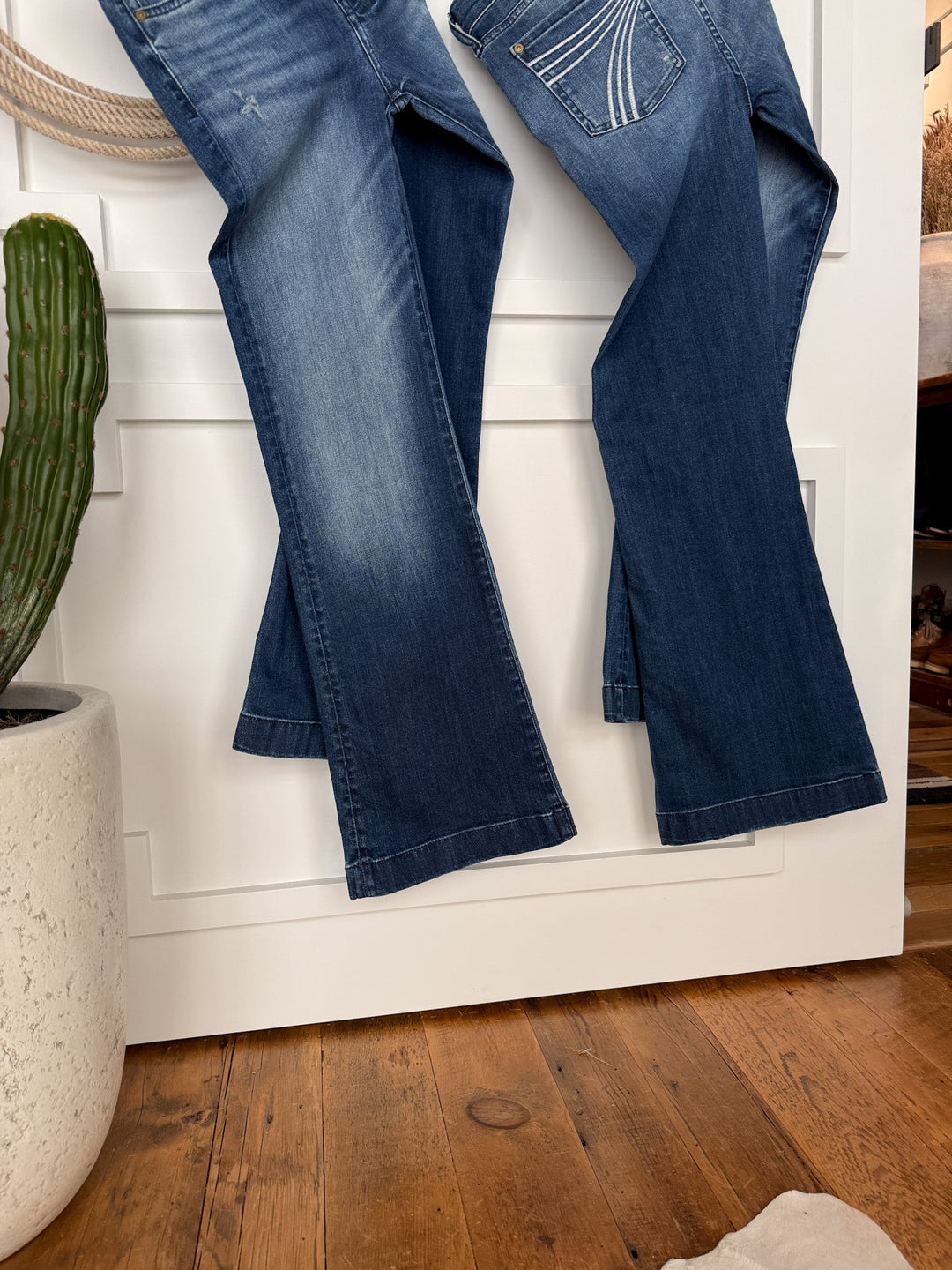 Two pairs of medium dark wash denim blue jeans, including The Beau 7 for all Mankind Dojo Trouser from Savannah Sevens western life{&}style, hang on a white paneled door beside a green potted cactus on a wood floor with a white textured rug.
