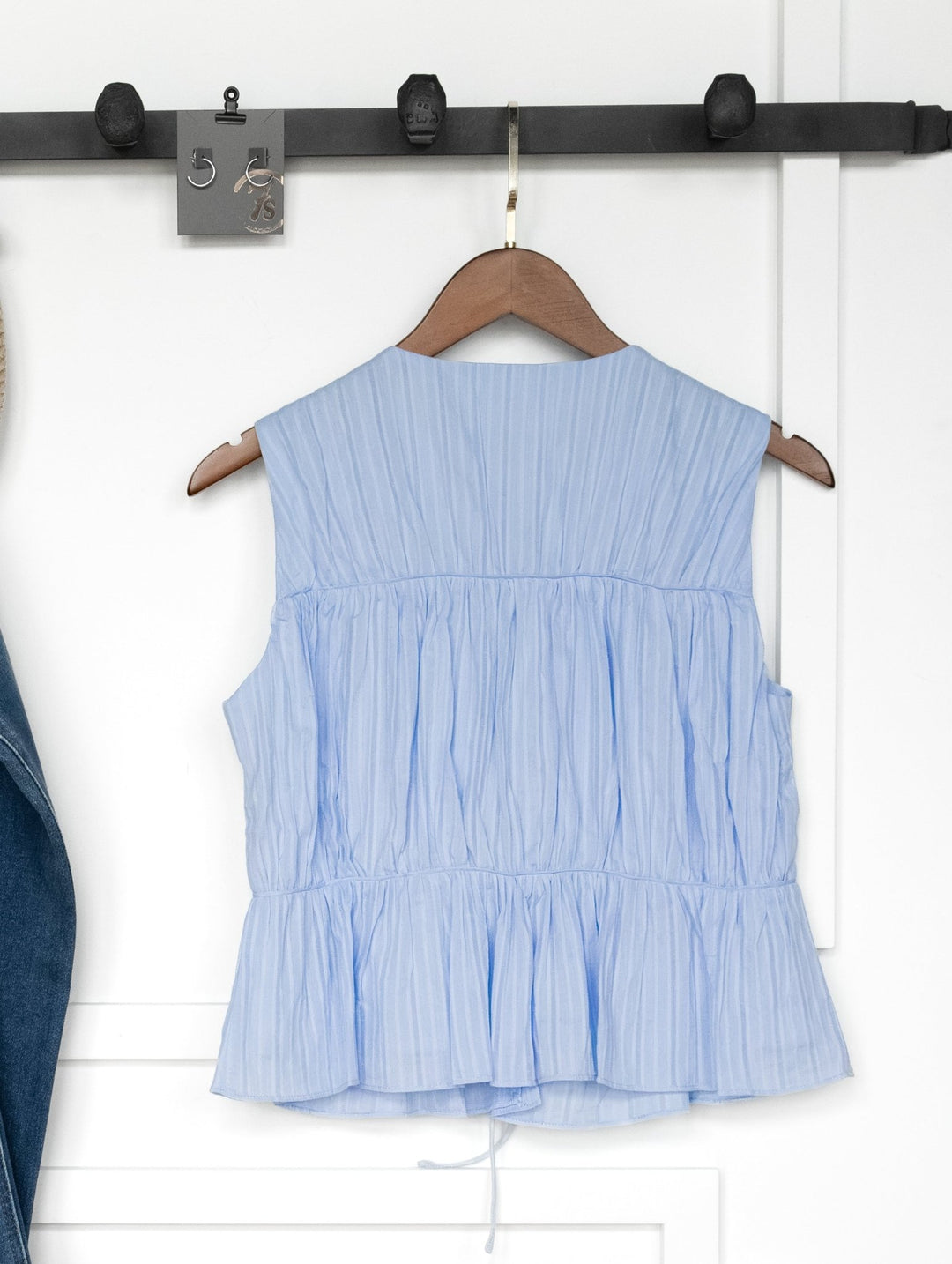 The Bella Villa Light Blue Tie Front Top by Savannah Sevens western life{&}style, a sleeveless cotton top with pleats, hangs on a wooden hanger from a black rack against a white wall; part of a denim garment appears on the left.