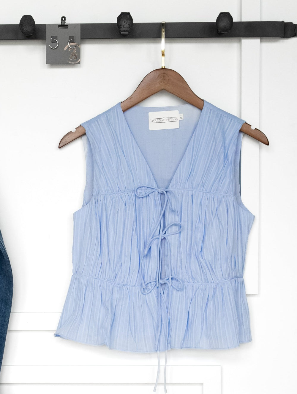 The Bella Villa Light Blue Tie Front Top by Savannah Sevens western life{&}style, a sleeveless cotton blouse with pleats and V-neck, hangs on a wooden hanger beneath a metal rack with hooks and keys against a white wall.