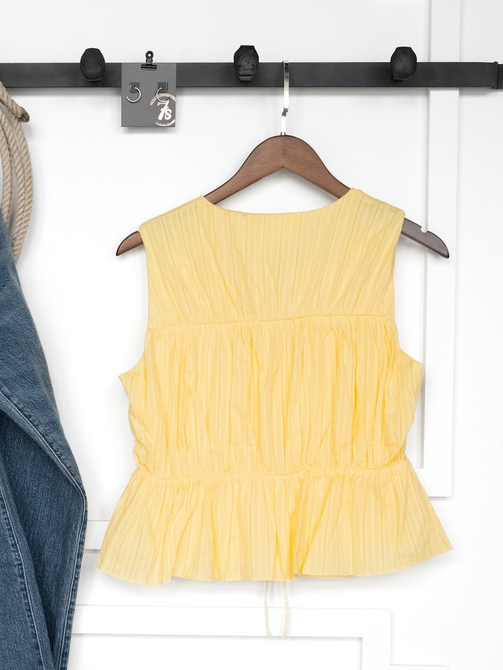 The Bella Villa Yellow Tie Front Top by Savannah Sevens western life{&}style, featuring a sleeveless design and ruffle hem, hangs on a wooden hanger against a white wall with part of a denim jacket visible on the left.