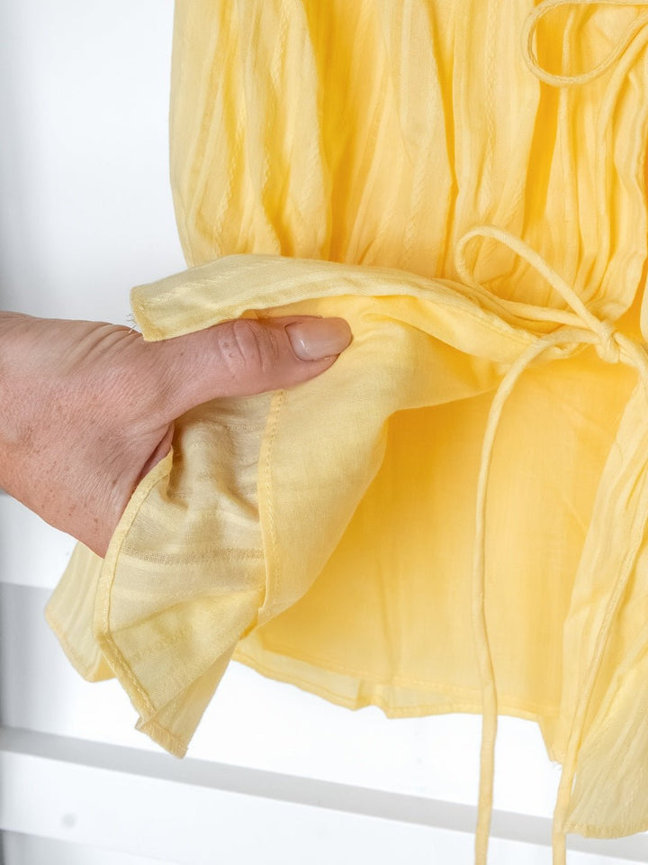 A hand holds the edge of the Bella Villa Yellow Tie Front Top by Savannah Sevens western life{&}style, a sheer piece with a gathered waist and delicate detailing.