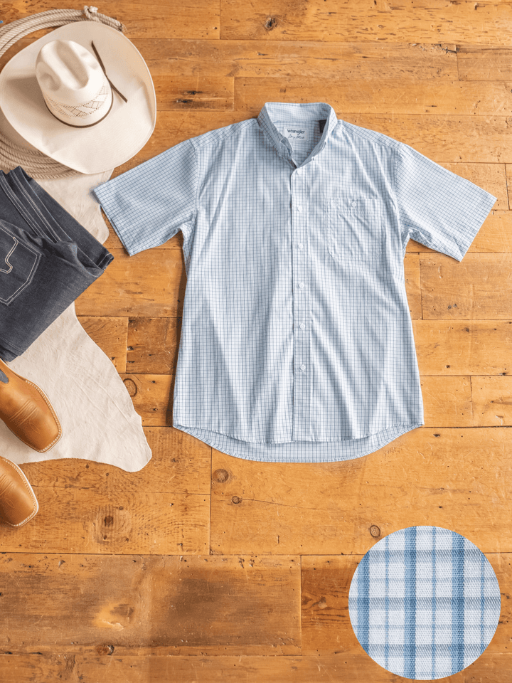 The Bentonville Wrangler Men's Blue Plaid Button Up from Savannah Sevens western life{&}style is laid flat on a wooden floor with a cowboy hat, jeans, and brown boots. A circular close-up shows the checkered fabric pattern.